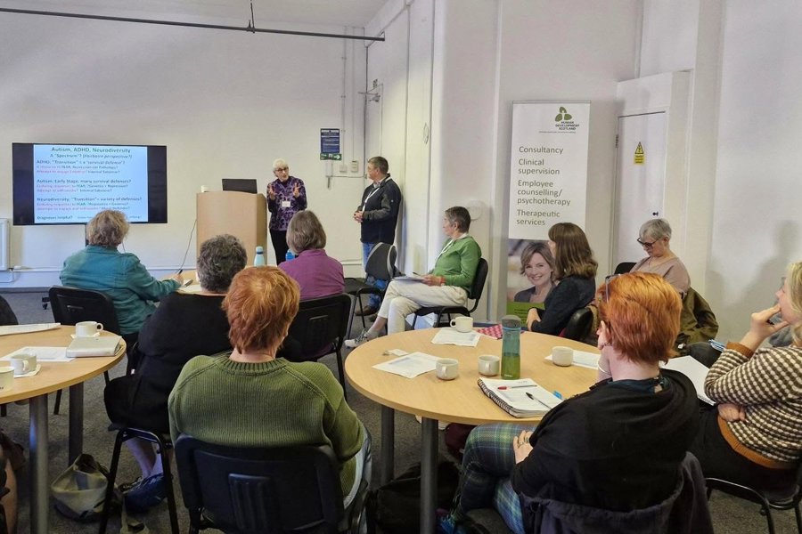 Two speakers present to UKCP members seated around tables in a training room in Glasgow, with a slide on autism, ADHD and neurodiversity on screen.