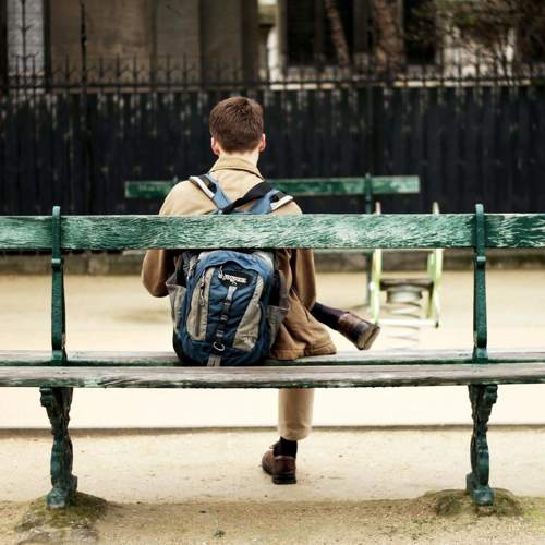 The back view of a young man with a backpack seated on the far side of a double-sided wooden bench